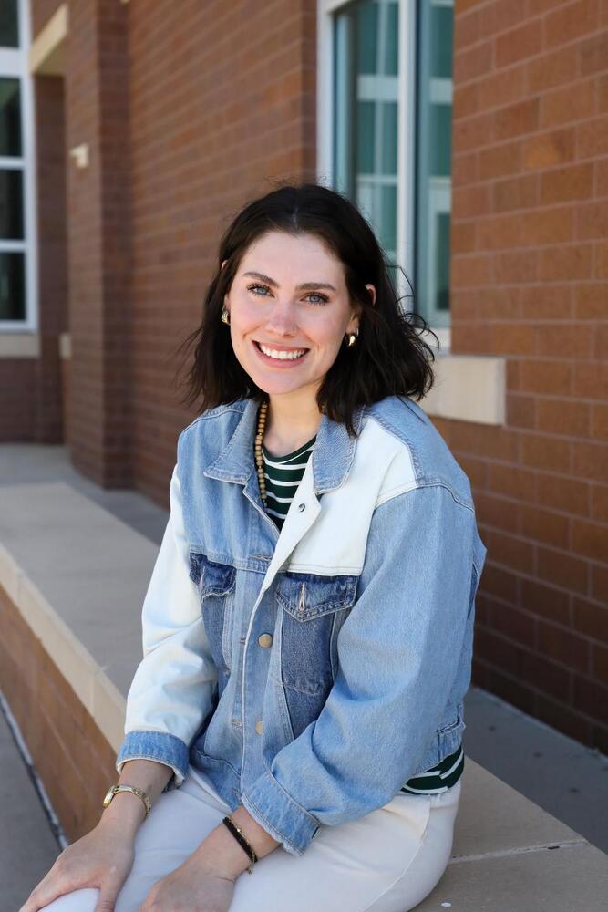 A woman, wearing a light-wash denim jacket, sits on a low wall outside a brick building. She smiles at the camera.