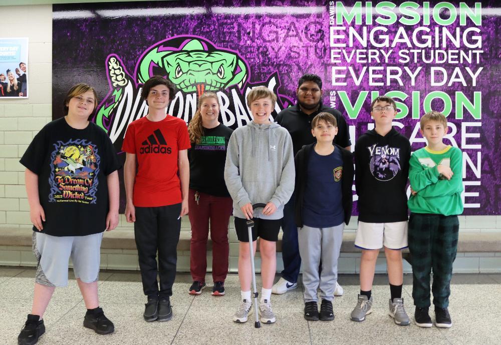 Eight students stand before a school mural. They are posing for a photo. The mural depicts a diamondback rattlesnake and text that includes "MISSION ENGAGING EVERY STUDENT EVERY DAY VISION".