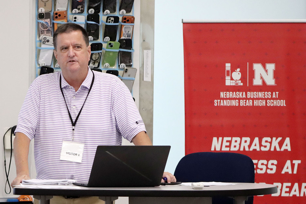 University of Nebraska-Lincoln professor Eric Thompson leads a lecture on the economic principles of demand during a Macro Economics class at Standing Bear High School. He is standing in front of a table that has his laptop computer on it. He is standing next to a banner that says "Nebraska Business at Standing Bear High School" with the logos of Lincoln Public Schools and UNL.