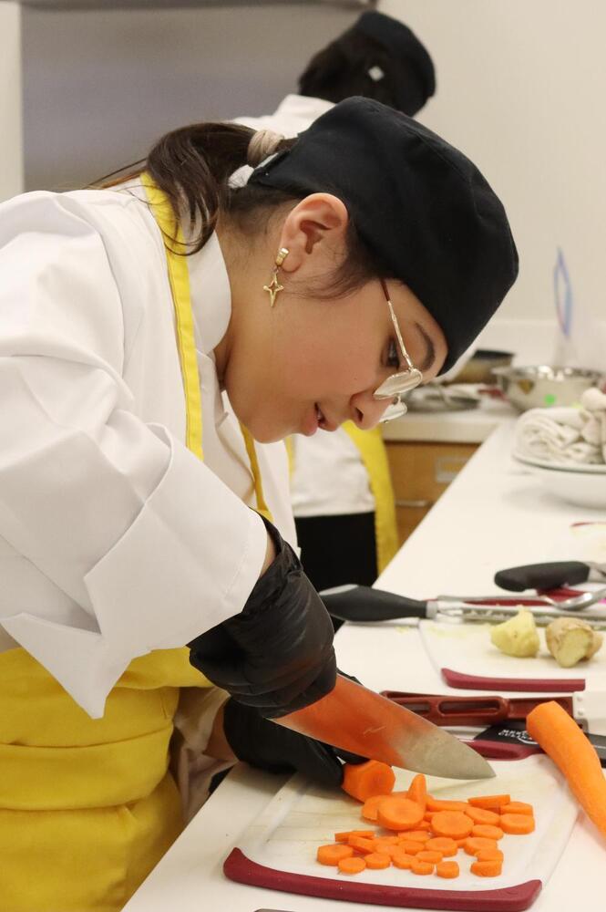 Southeast student Odulia slices a carrot as part of the team entree competition at the LPS Culinary Contest. She is wearing latex gloves on both hands. She is using her right hand to position her knife over the carrot and is using her left hand to hold the carrot in place. She has already cut more than a dozen pieces on the cutting board. Odulia is wearing glasses and a culinary cap over her hair.