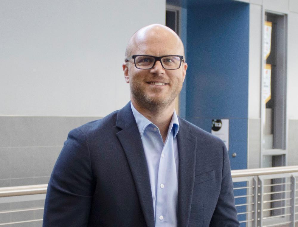 The Career Academy Director Josh Jones smiles on the second floor of the TCA building while wearing a blue collared shirt.