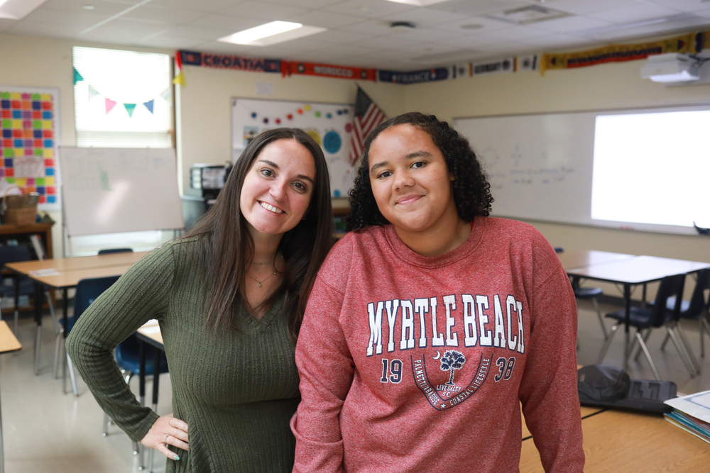 Two people smile for a photo in a classroom, standing side by side in front of desks, whiteboards and a projector screen.