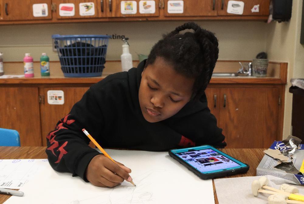 A young person with braided hair, wearing a black hoodie with red sleeves, intently sketches on white paper with a pencil. A tablet displaying images sits nearby on the wooden table in what appears to be a craft room.