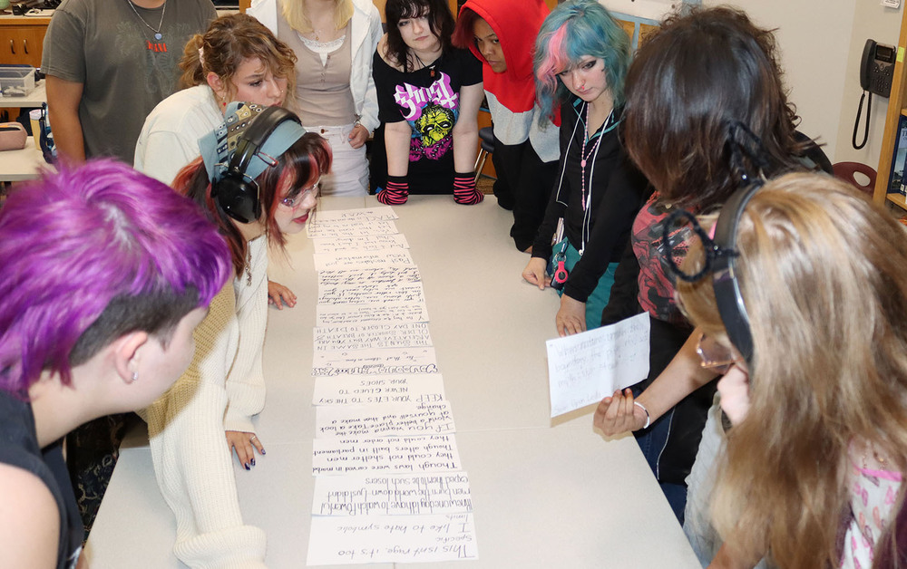 Ten Arts and Humanities students gather around a table to work on a lyrics project in their classroom. They have lined up small pieces of paper with song lyrics on them. They are deciding what order to place the pieces of paper in to create a new song.