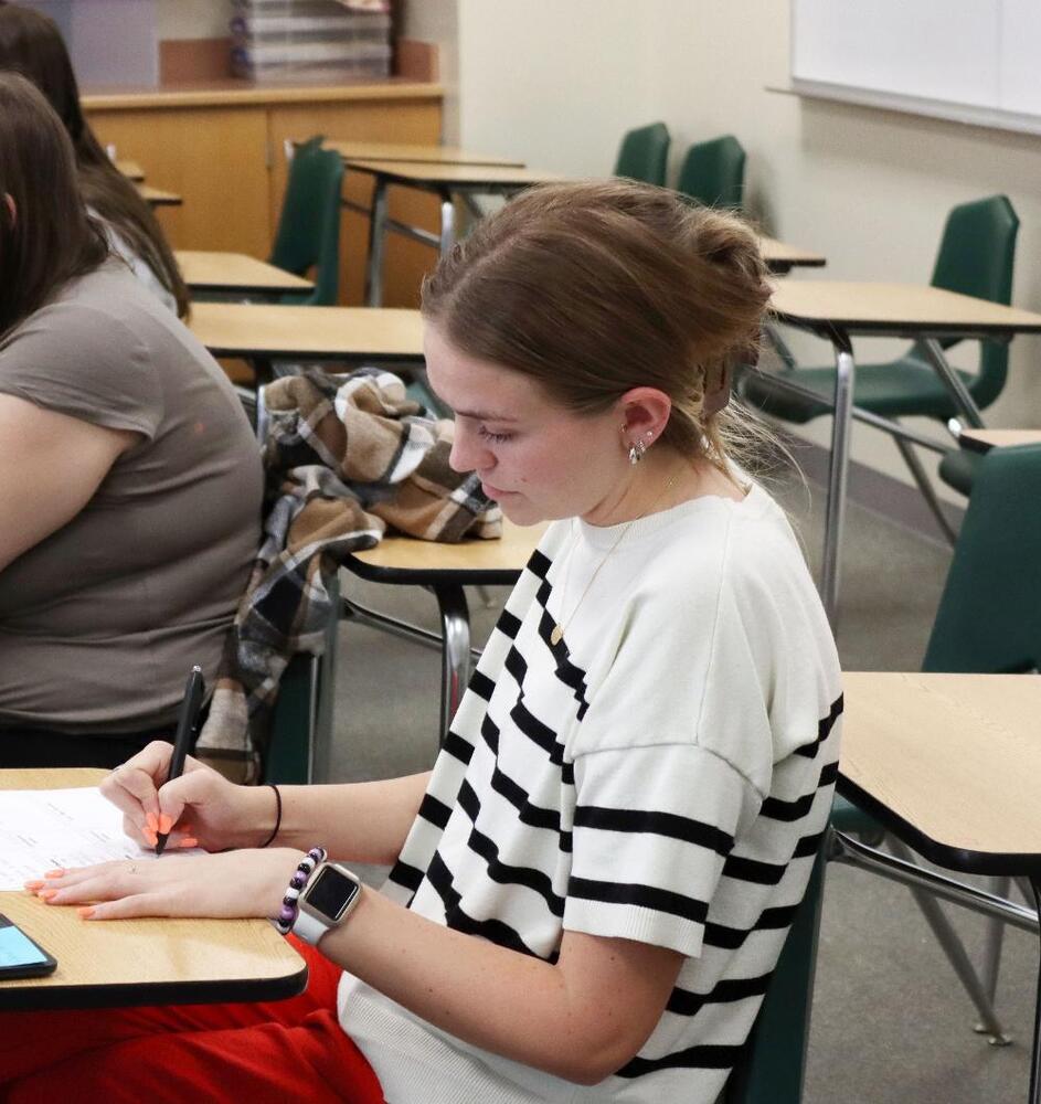 A female teacher is sitting at a desk in a classroom at Southwest High School for the LPS ElevatED professional learning program. She is holding a pen with her right hand to write answers to a question on a piece of paper. She has a watch on her left hand and is wearing a shortsleeved shirt.