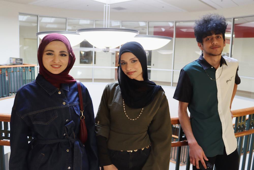 Three smiling young adults, two women in hijabs and a man, stand behind an indoor wooden railing. One woman wears a burgundy hijab and denim jacket, the other a black hijab and olive top. The man wears a green, black, and white split-color shirt.