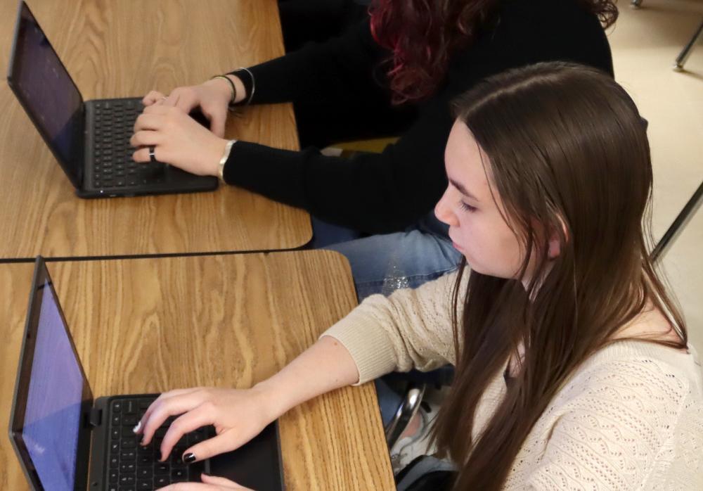A Southeast High School student is typing on her laptop computer during a newspaper class this fall. She is facing right to left and has both hands on her keyboard. She is wearing a light sweater and has her sleeves rolled up on her right arm. The hands of another student typing on a second keyboard are shown at the very top of the picture.
