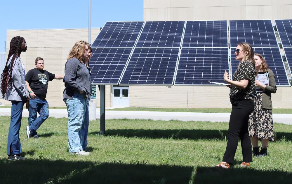 Four people stand near a solar panel array. Two women discuss, holding papers. A man and woman observe. The setting is a grassy area outside a building.