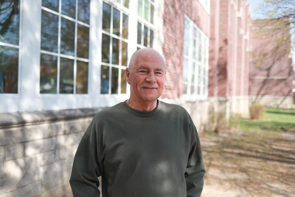 A bald, smiling man in a dark green shirt stands outdoors. Behind him is a red brick building with white-framed windows showing tree reflections, and green grass visible on the right.