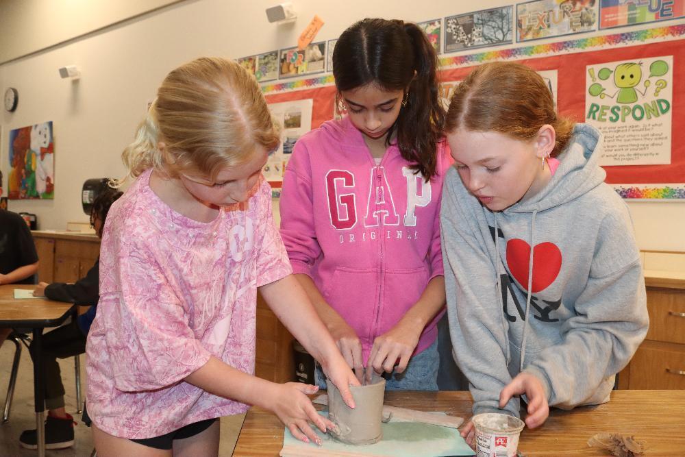 Three young girls intently shape clay together at a wooden table in an art classroom. The girl on the left forms a cylindrical object, while her friends assist, all focused on their hands-on creative project.