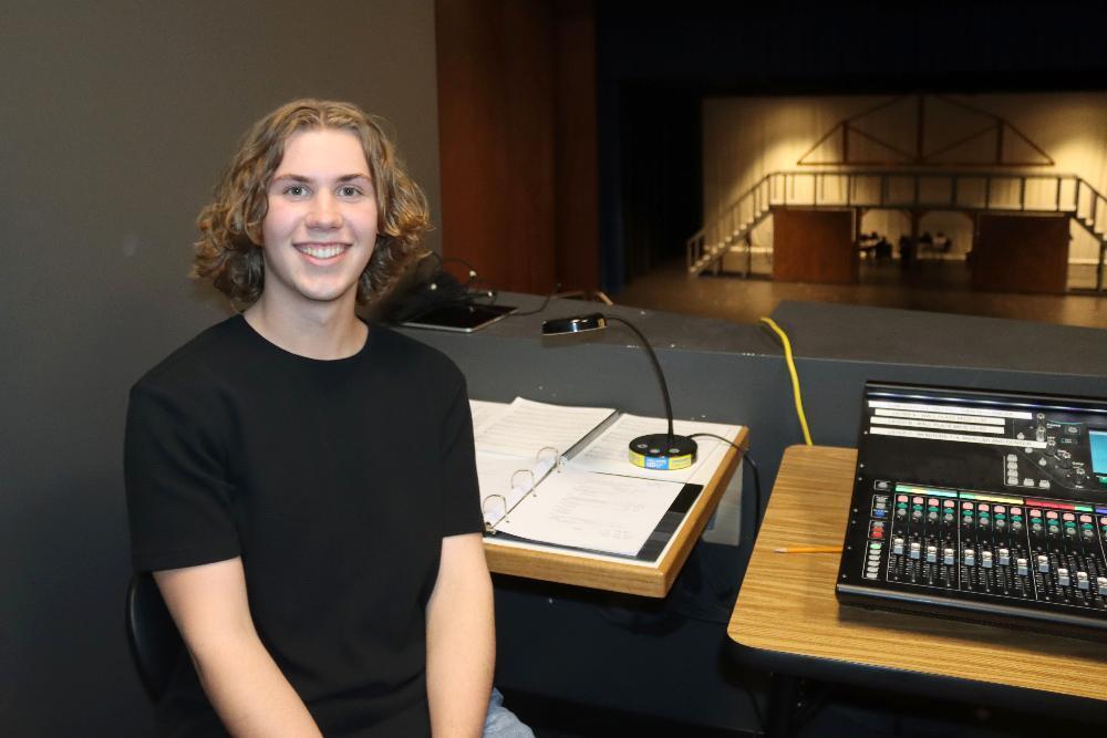 A smiling young man with curly hair sits in a theater control booth. Behind him, a sound mixing board and script are visible. Further back, a stage set features wooden stairs and a bridge, ready for a production.