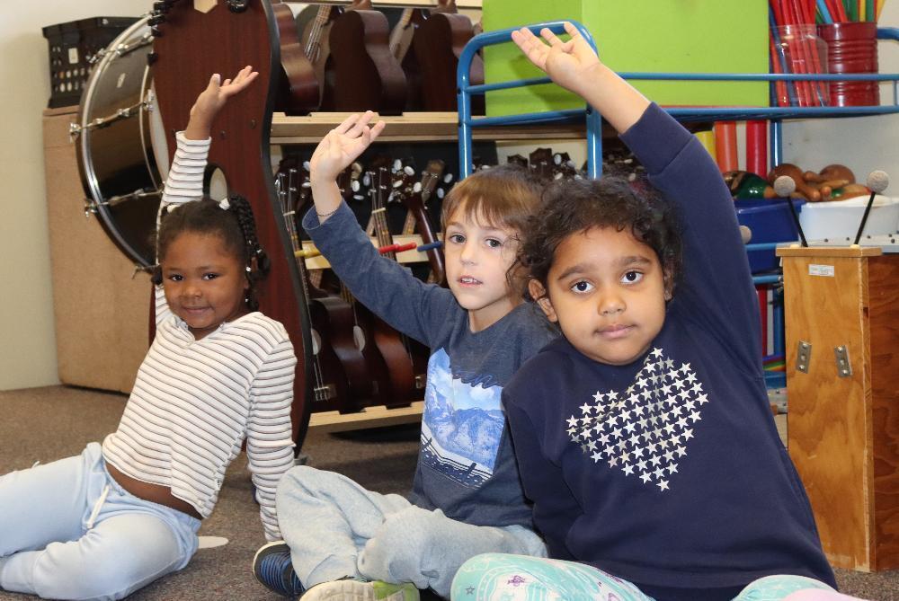 Three Eastridge Elementary School students smile in a music classroom this fall. They are raising their hands to indicate that they are displaying good effort in class. A girl at the far lefthand side of the photo is raising her right hand, a boy in the middle is raising his right hand and a girl on the righthand side is raising her left hand. They are sitting in a row in front of several musical instruments.