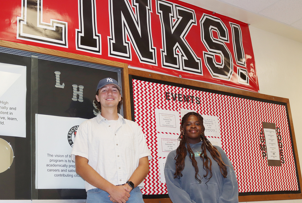 From left, International Baccalaureate students Everett and Marione stand next to each other in front of a school bulletin board. There is a large poster that say, 'Links!' above them and pieces of paper with events listed on the bulletin board.