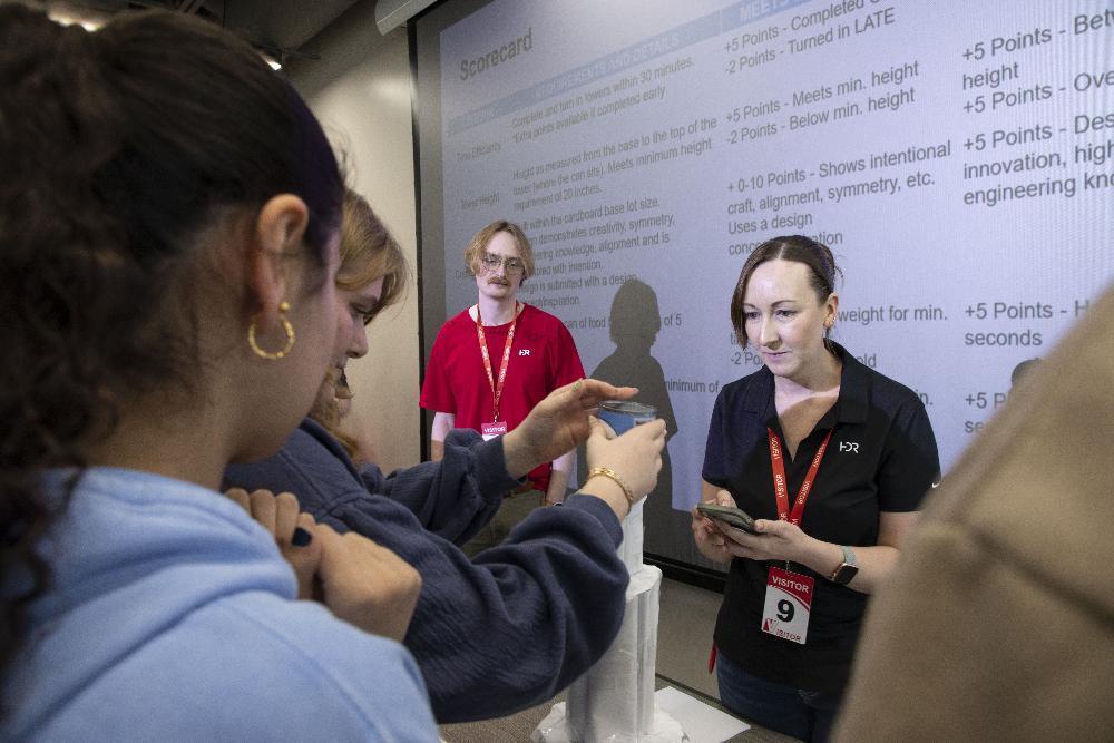 People surround a white tower. One places an object atop it, as a man in red and woman in black with a phone observe. A "Scorecard" outlining building challenge rules is projected behind them.