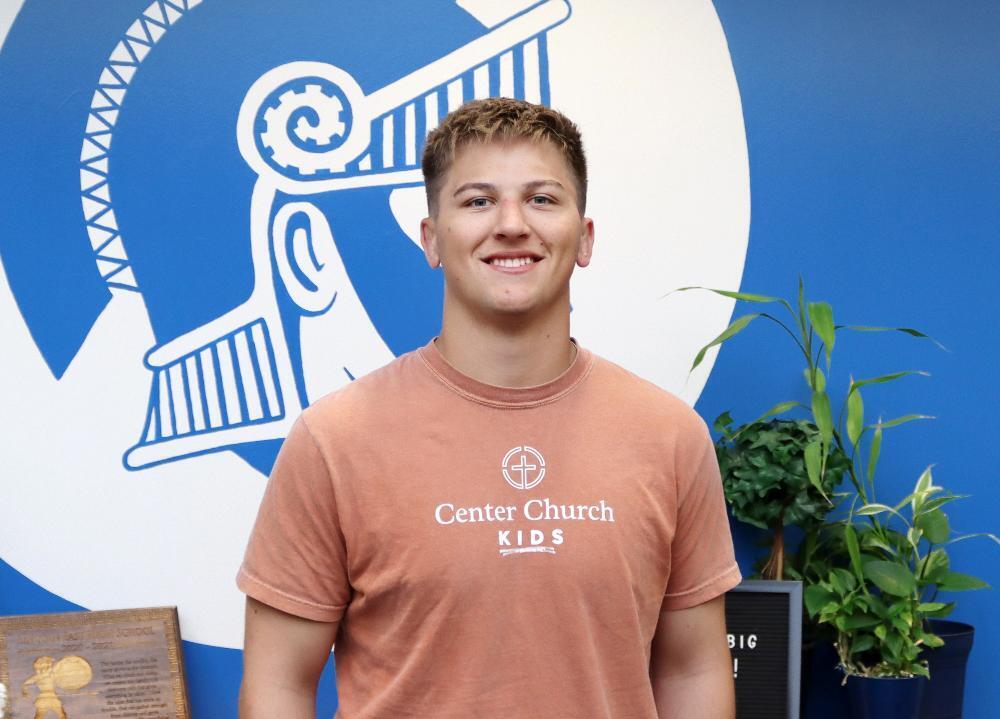 A young man smiles, wearing a peach "Center Church Kids" t-shirt, standing in front of a blue wall with a stylized helmet graphic.