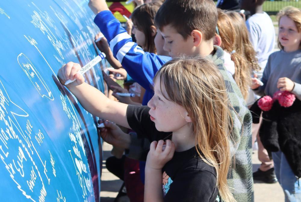 A girl uses a pen in her right hand to sign her name to a large pipe at the intersection of 98th Street and White Pine Road. She is in the middle of a large row of students who are signing their names on the pipe with markers and pens. The activity was part of a field trip that taught students about water and engineering.
