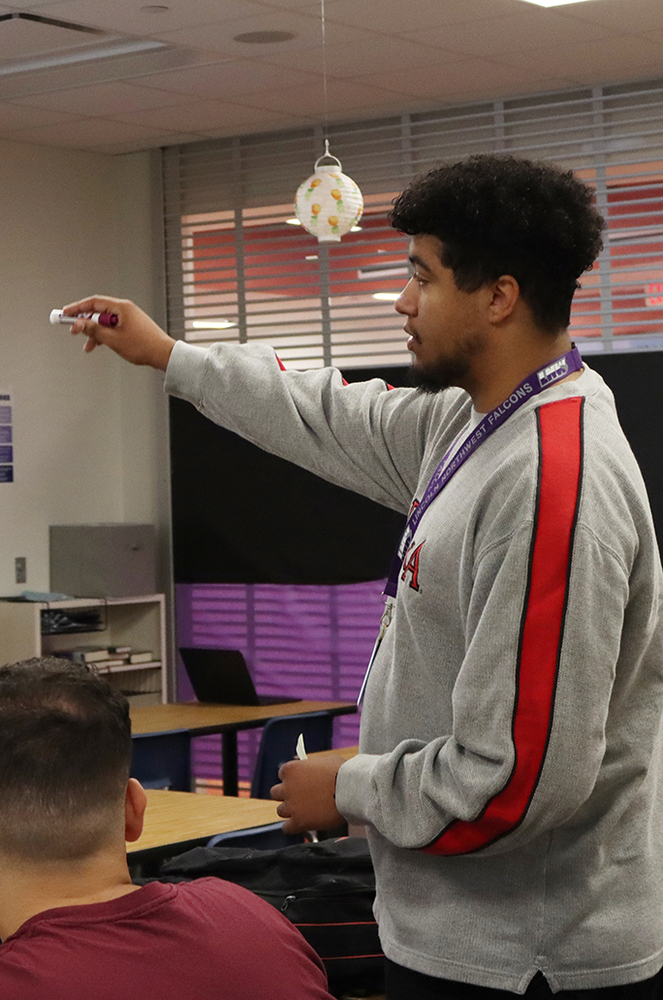 Northwest teacher Isaiah Collier is standing right to left in a classroom. He is holding a round eraser in his right hand and is pointing towards a monitor at the front of the room.