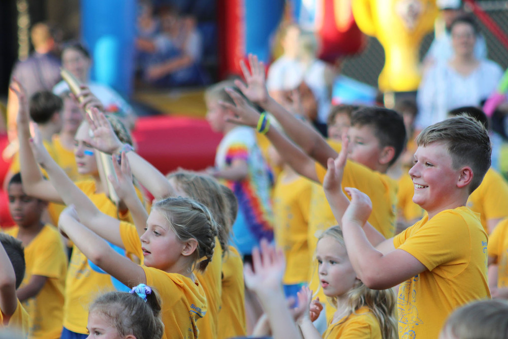 More than a dozen Sheridan Elementary School students smile as they take part in a Dancing Through the Decades activity. They are facing right to left and are standing in a grassy field outside the school building.