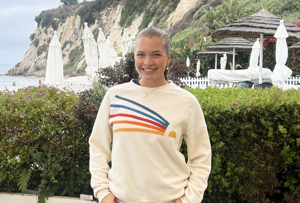 East student Avery Nordhues smiles as she stands in front of a hedge of bushes at a restaurant on the Pacific Coast. Tall sandy cliffs are in the background by the Pacific Ocean.