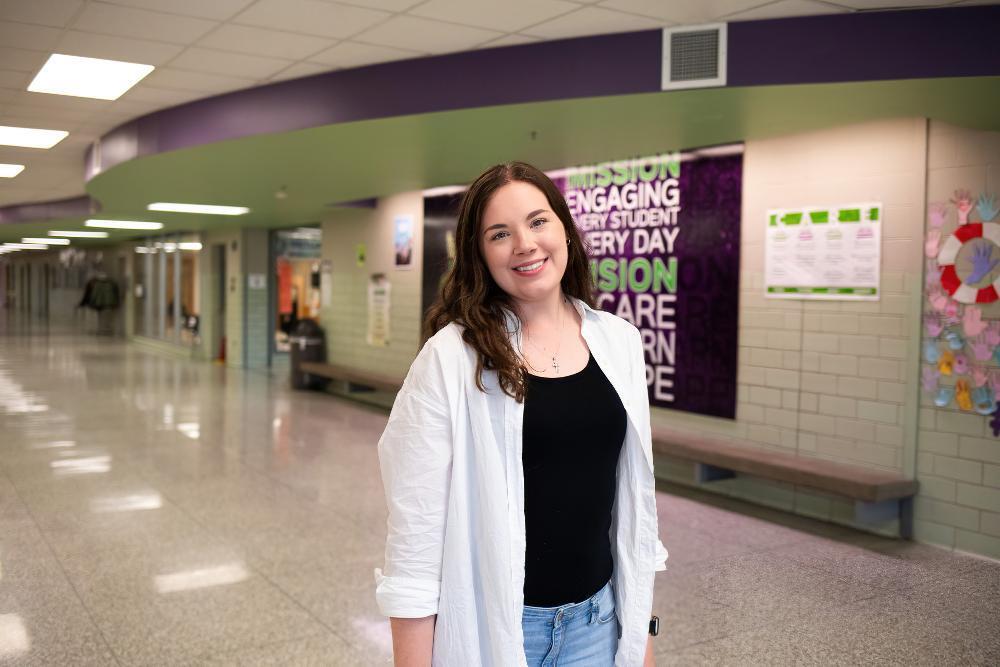 A young woman stands smiling in a school hallway. She wears a white shirt over a black top and jeans. A wall poster reads: "MISSION ENGAGING EVERY STUDENT EVERY DAY VISION CARE LEARN HOPE."