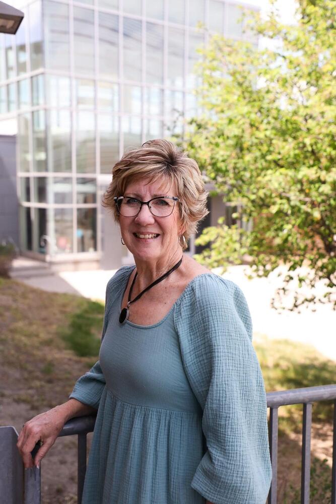 A woman wearing glasses and a teal dress leans on a railing. She smiles at the viewer. The background includes a modern building and a tree.