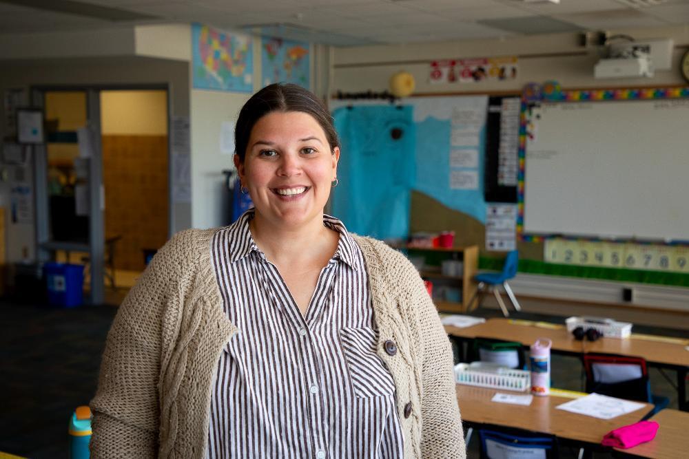 A woman standing in a classroom wearing a cardigan and striped shirt.