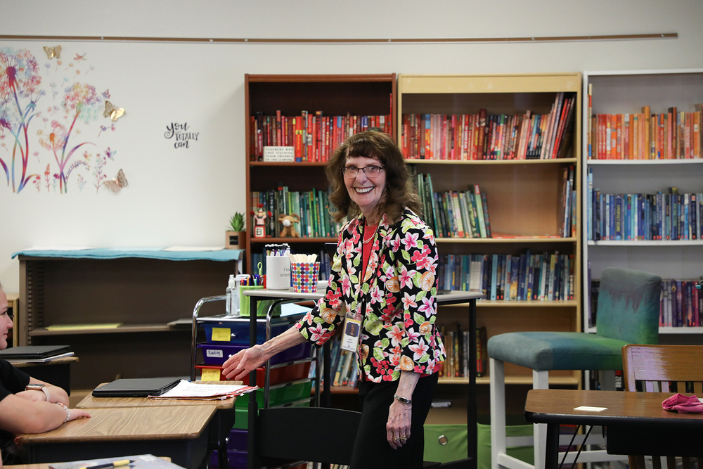 Diane Huseman smiles as she teaches in a Morley Elementary School classroom. There are bookshelves behind her and a student is smiling at a desk next to her.