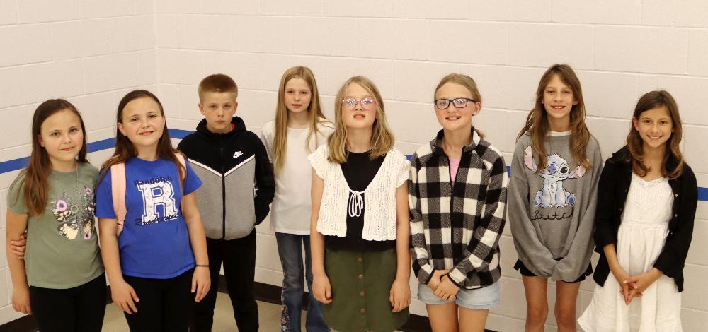 Eight children stand against a white-tiled wall wearing various casual clothes at Randolph Elementary School.