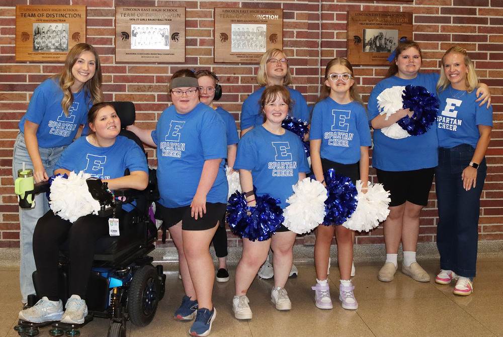 East inclusive cheer team coaches Anna Schulz and Sydney Mickells stand with seven cheerleaders in a school lobby. They are standing in a row in front of a brick wall with plaques on it.