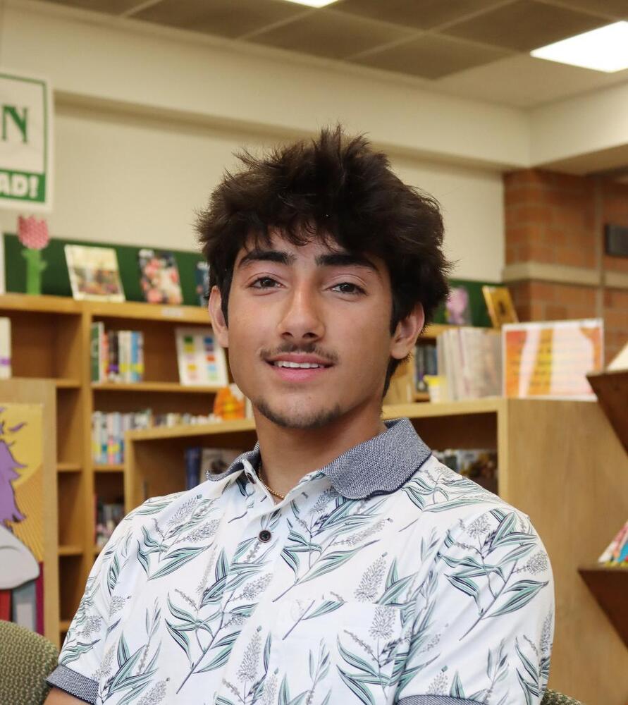 A young man with dark hair, wearing a white, leaf-patterned polo shirt, sits in a library.