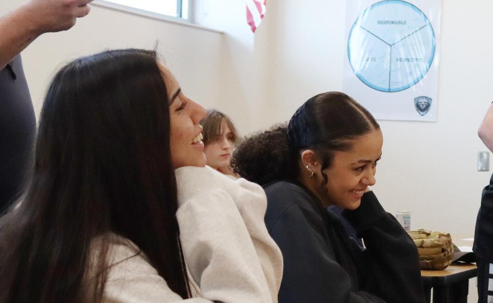 Two young women laugh in a classroom. One wears a light beige sweatshirt; the other, dark clothing. A partially visible chart depicting "Responsible," "Fair," and "Respectful" is on the wall.