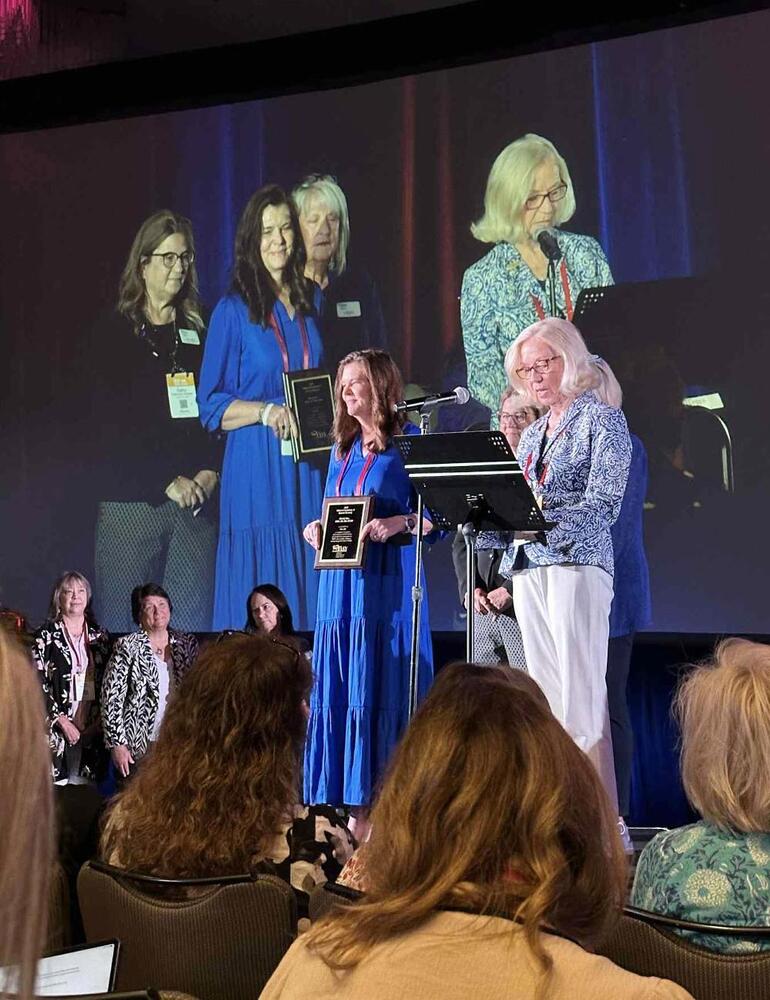 LPS Director of Health Services Wendy Rau stands on a stage and holds a plaque that recognizes her as a National Association of School Nurses School Nurse of the Year.