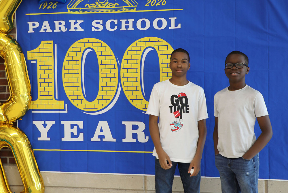 Two Park Middle School students stand in front of a banner during the 100th anniversary of the school building. The banner says 'Park School - 100 Years - 1926-2026' and is hanging next to three balloons spelling out the numeral 100.