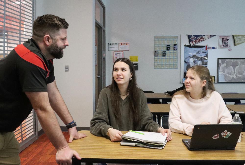 A male teacher addresses two female students seated at a desk in a classroom. They are working on schoolwork; papers and a laptop are present.