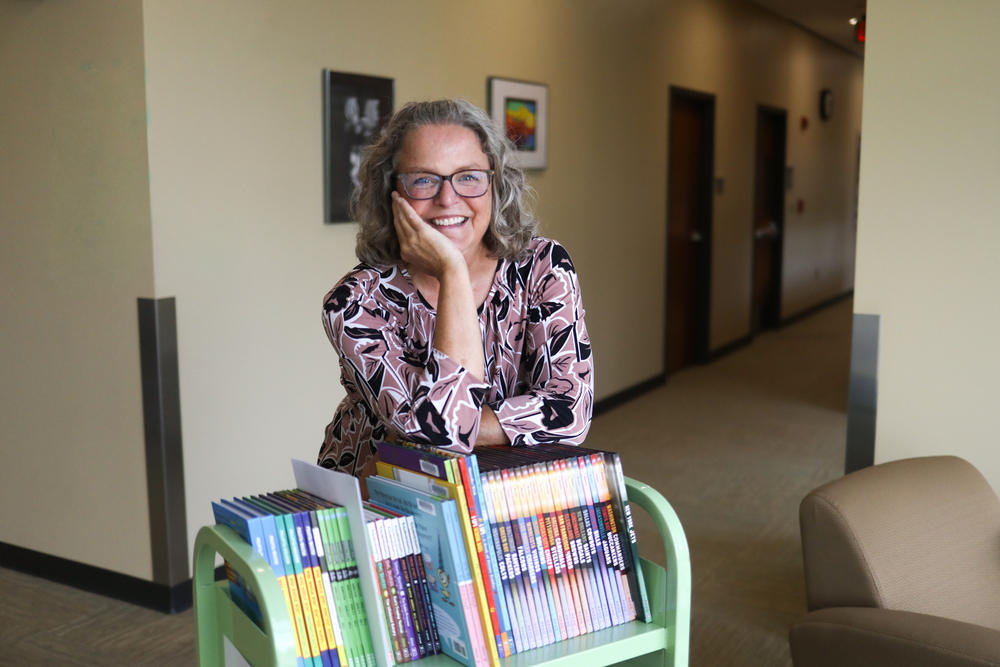 Woman resting elbow on a rack books. She is smiling wearing glasses.