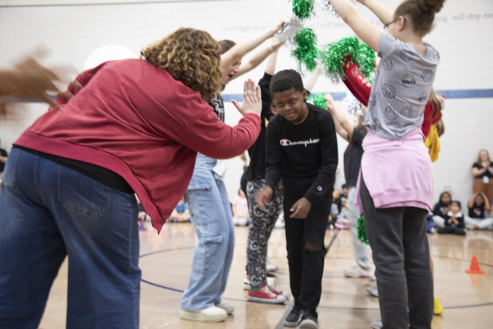 A woman in a red jacket high-fives a boy in a black sweatshirt. Children are forming an arch overhead with green pom-poms in a gymnasium.