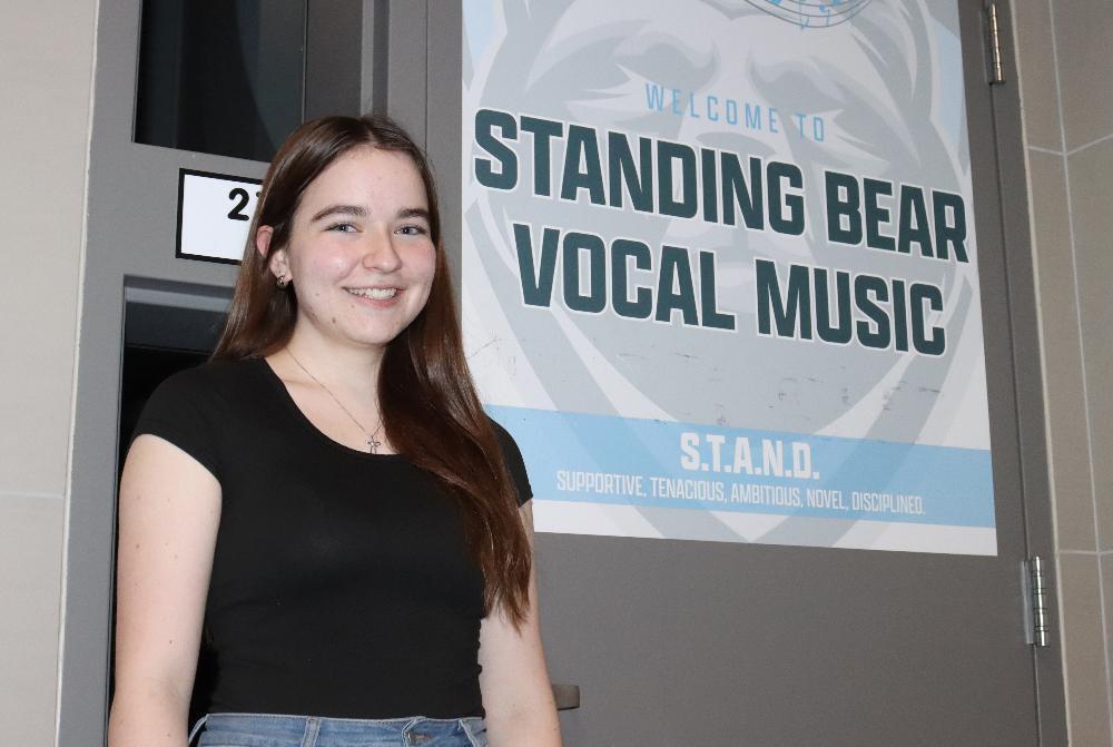 A smiling young woman with long brown hair stands in a hallway. Behind her, a door displays a banner saying "WELCOME TO STANDING BEAR VOCAL MUSIC." She wears a black top and jeans, next to a door labeled "2".