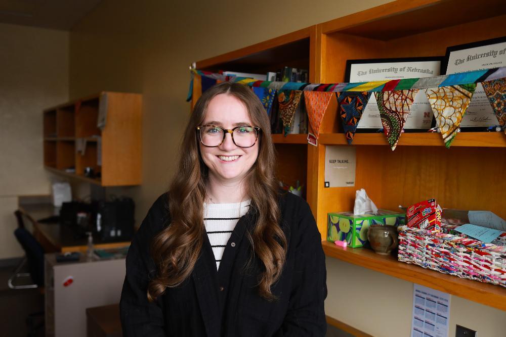Smiling woman with long brown hair and glasses standing in an office, with wooden shelves behind her holding books, framed diplomas, colorful fabric bunting and office supplies.