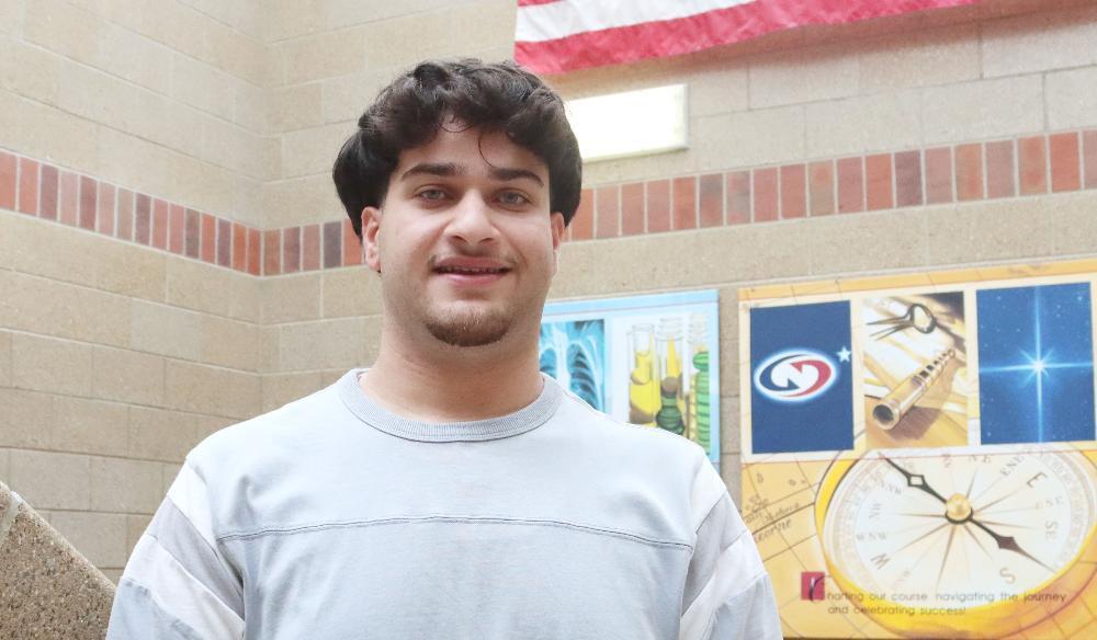 A young man with dark, curly hair and a goatee smiles slightly, looking at the camera. He wears a light grey shirt. Behind him are a brick wall, part of an American flag, and educational posters featuring science imagery, a compass, and stars.