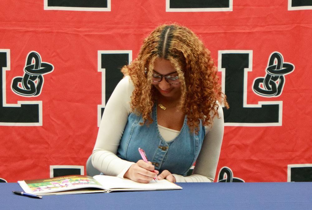 A young woman with curly auburn hair and glasses smiles while signing an open book with a pink pen. She's at a blue table, with a red banner featuring black 'L's behind her.