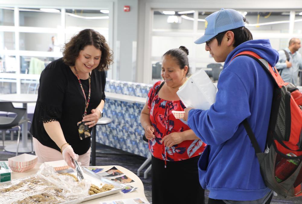 A woman offers cookies from a tray using tongs to two other people in a bright, modern building.