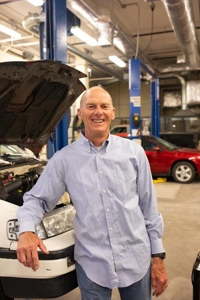 A man leans on a car with its hood up, smiling in an auto repair shop.