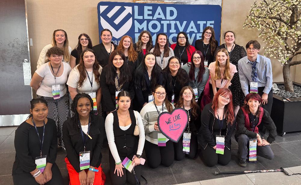 A diverse group of smiling young adults, mostly women, pose for a photo. They wear lanyards, some kneeling, against a blue "LEAD MOTIVATE ACHIEVE" banner. One holds a pink "WE LOVE Ed Tech" heart sign. A cane rests on the floor.