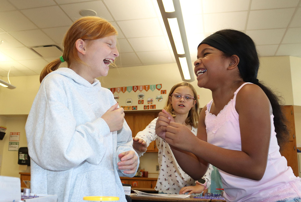 Two Kloefkorn Elementary School girls laugh with each other during a snap circuits activity in their fourth grade classroom. A third girl is smiling as she prepares to tap one of the girls on her arm.
