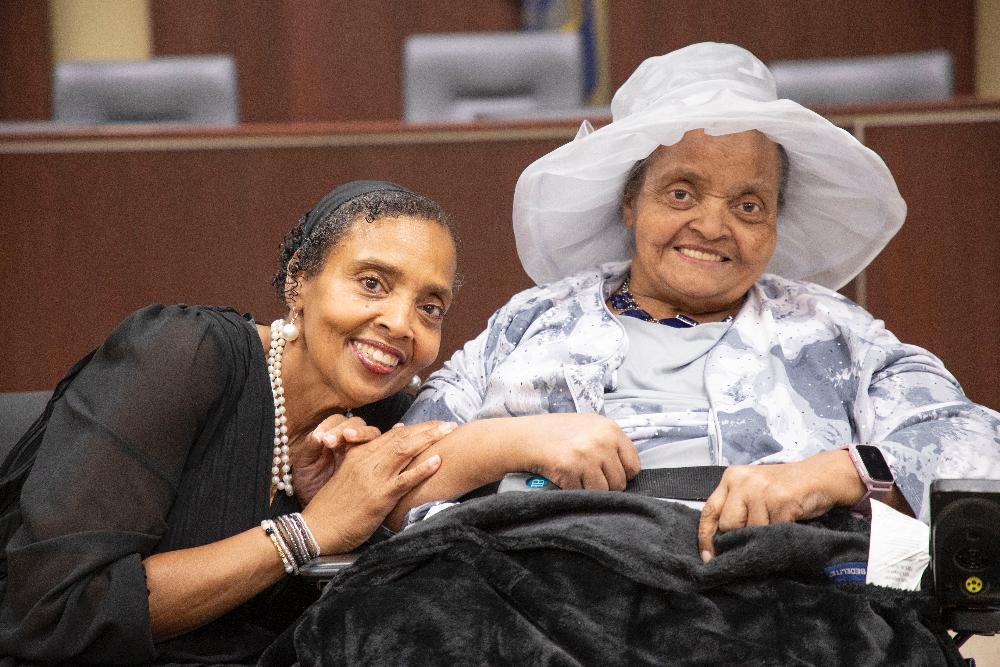 Two women sit together. One, wearing a hat and light gray jacket, is seated in a wheelchair. The other woman leans in, holding her hands. The setting appears to be a formal hall.