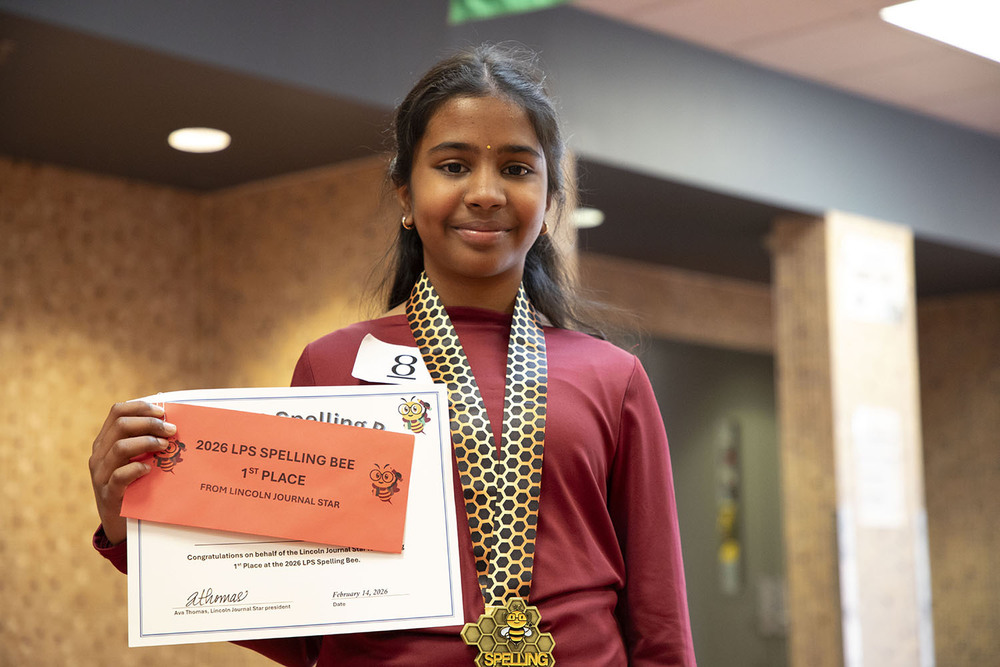 2026 LPS Spelling Bee champion Hanshiya smiles with her first-place certificate. She is wearing a championship medal around her neck and smiling in a school hallway.