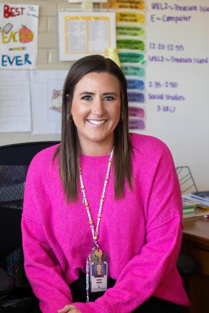 A woman wearing a pink sweater and lanyard smiles. The lanyard displays her name, "Katelyn Johnson," and a school ID. The background shows a classroom with educational charts.