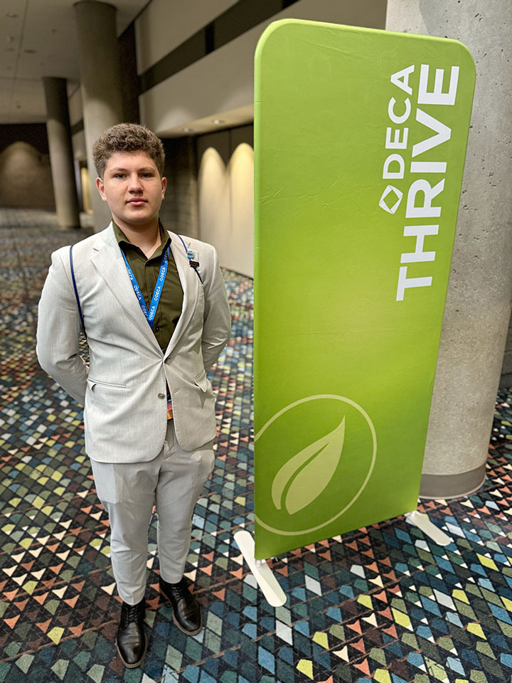 Southwest student Ruvim Orlov stands next to a large rectangle-shaped sign at the DECA International Career Development Conference. The sign has a symbol of a leaf and the words DECA Thrive on it.