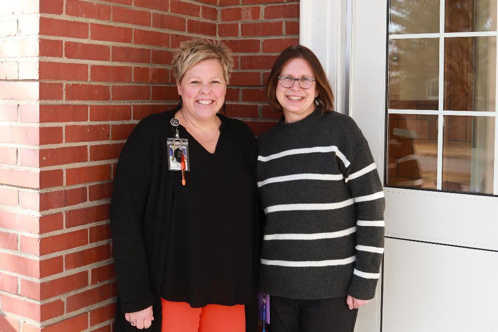 Two smiling women stand side-by-side. The woman on the left has short blonde hair and an ID badge. The woman on the right wears glasses and a dark striped sweater. They are in front of a brick wall and a white door with windows.
