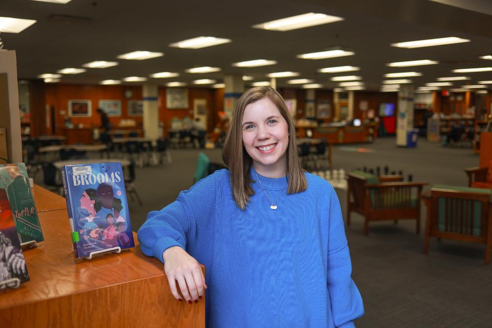 A woman standing inside a library wearing a blue sweater.